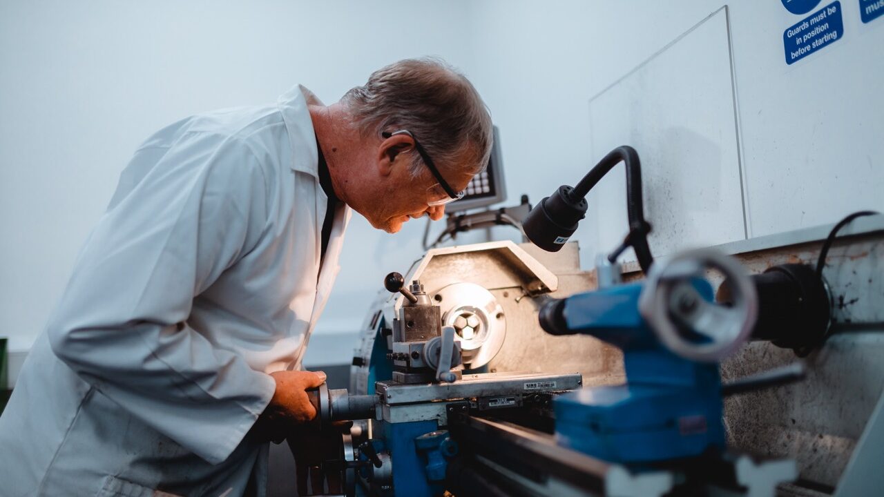 A man wearing a white medical coat adjusts an illuminated machine in a workshop