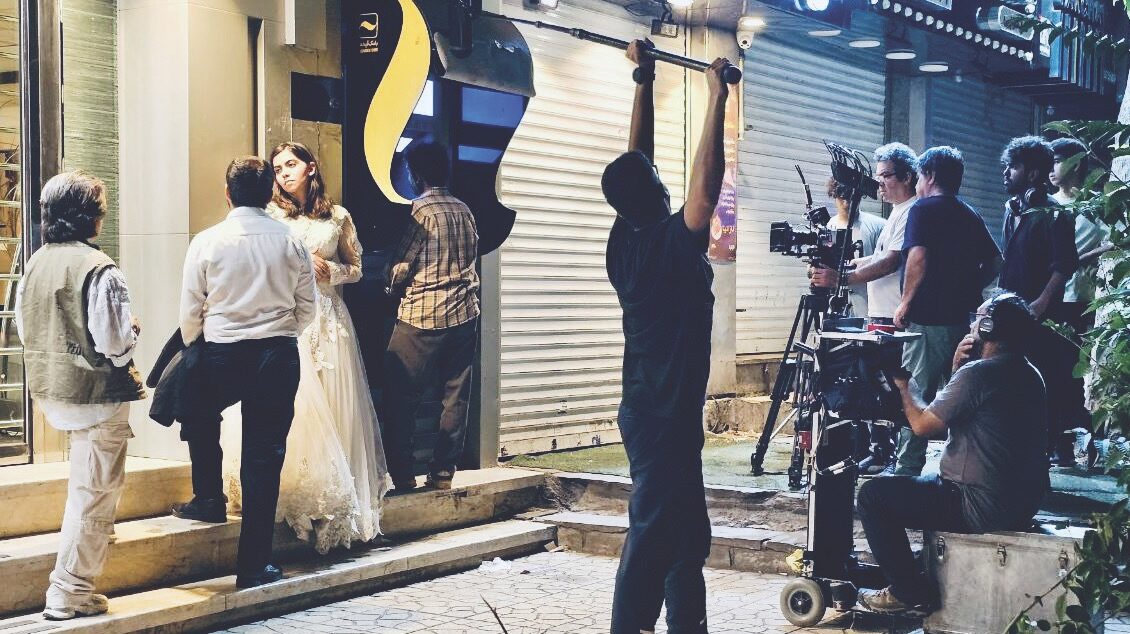 A bride and a groom stand outside a shop front, next to a person using an ATM. A camera crew records the scene