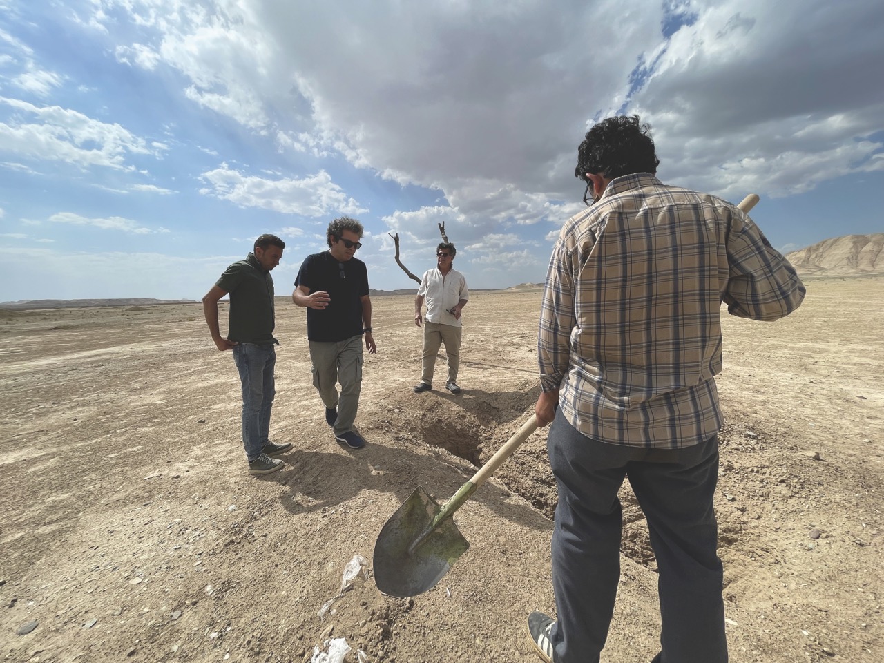 A man with a shovel stands in the foreground while three men direct him. The group stands in a flat desert expanse