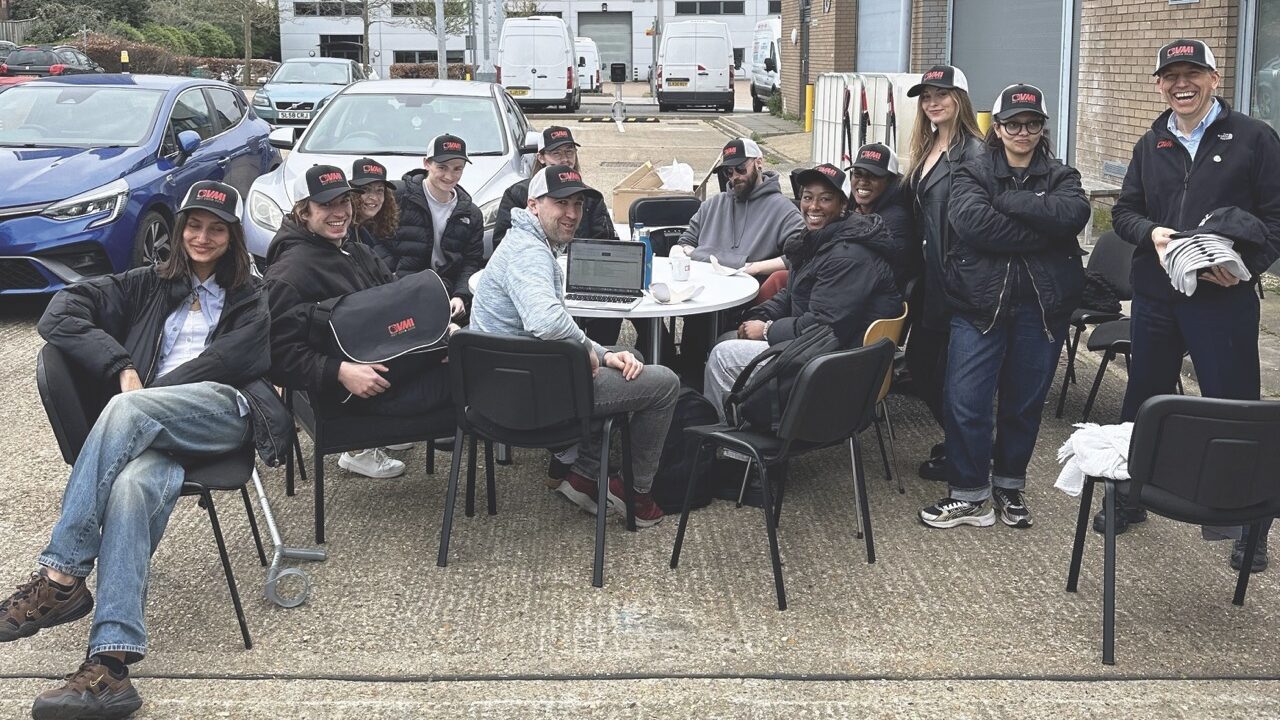 A big group of people wearing matching hats sit around a round table outside in a car park