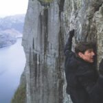 A man wearing black climbing up the sheer face of a mountain with a large lake below
