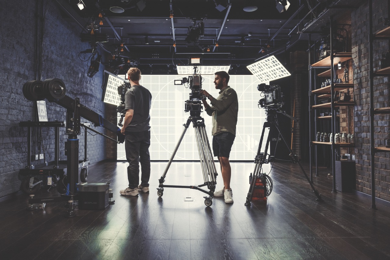A group of men stand next to a large camera and film equipment. They are backlit from a large LED wall behind