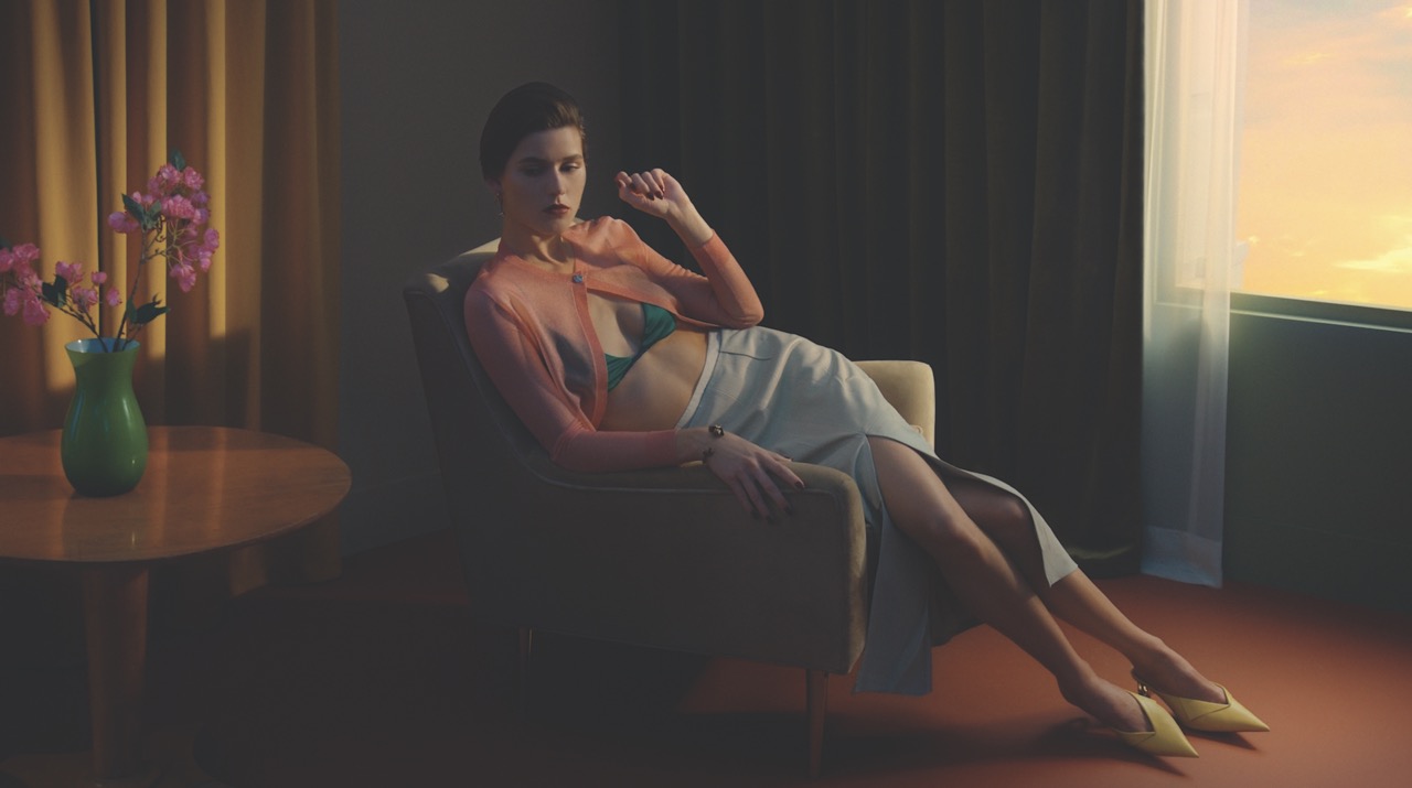 A woman wearing a skirts and sheer top lounges on an arm chair in a sunset lit room