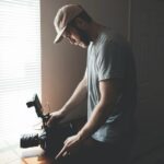 A man standing near a window wearing a cap and filming low down with a professional camera