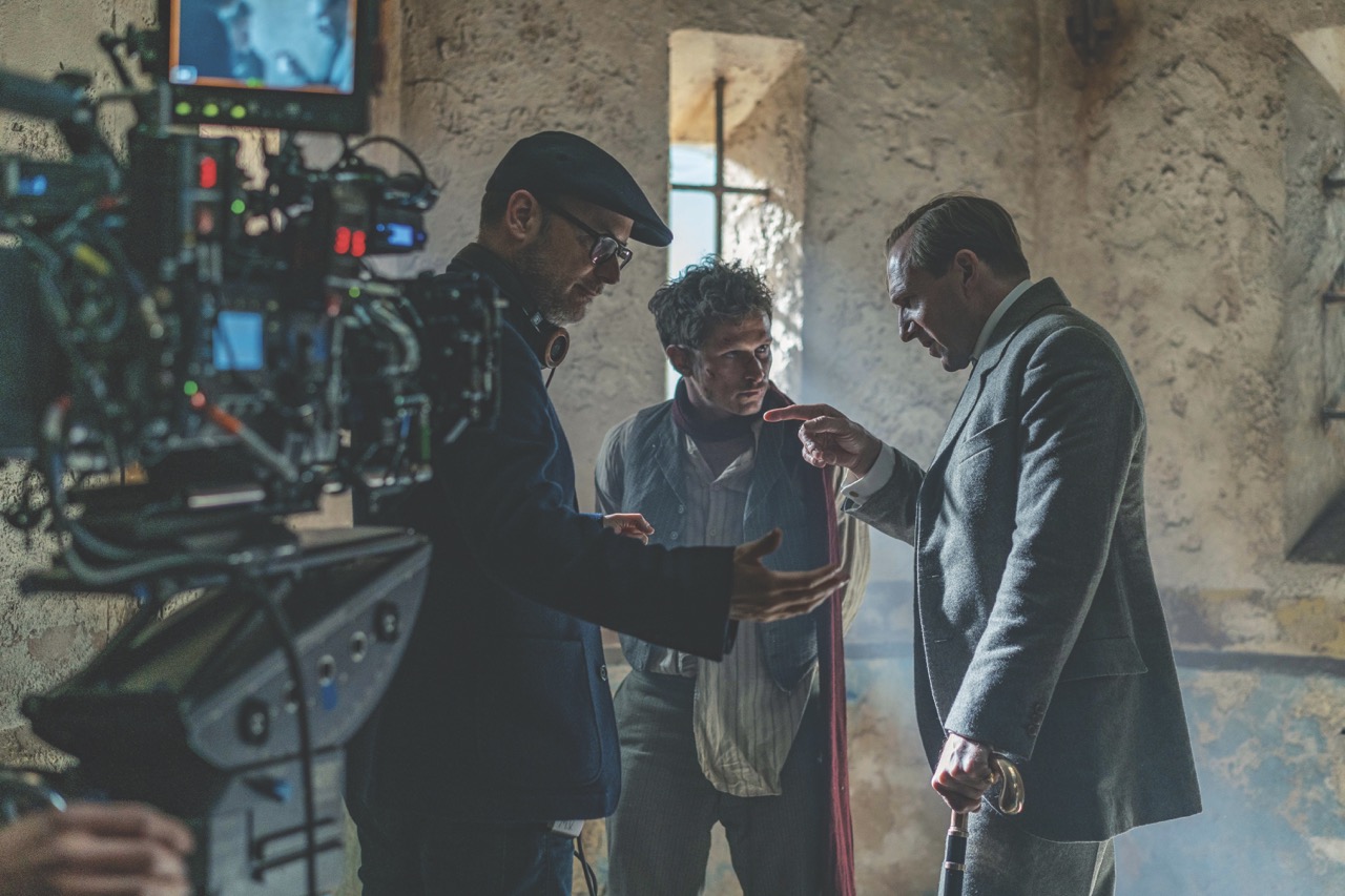 Three men stand and discuss in a circle with a camera in the blurred foreground