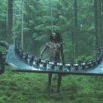 A zombie with long hair walks through a dense forest while being films by a rig of iPhones. This rig is carries by three men