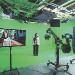 A woman stands in front of a green screen being filmed by a large camera. A monitor shows the same woman on a street