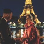A man and a woman stand opposite each other smiling. They stand next to a river in front of the Eiffel Tower at night