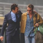 Two men walk down a street smiling with their arms around each other. One man wears a suit while the other carries a duffle bag and wears a brown jacket and sunglasses