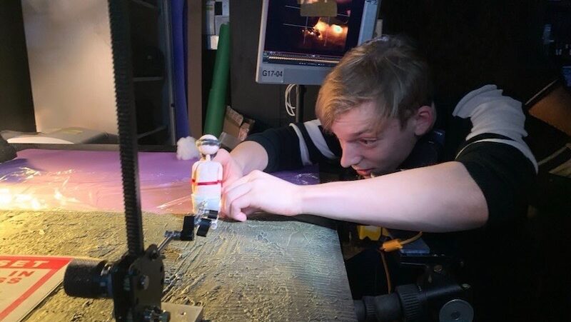 A man kneels and adjusts a figurine under studio lighting on a table. He create a stop motion animation