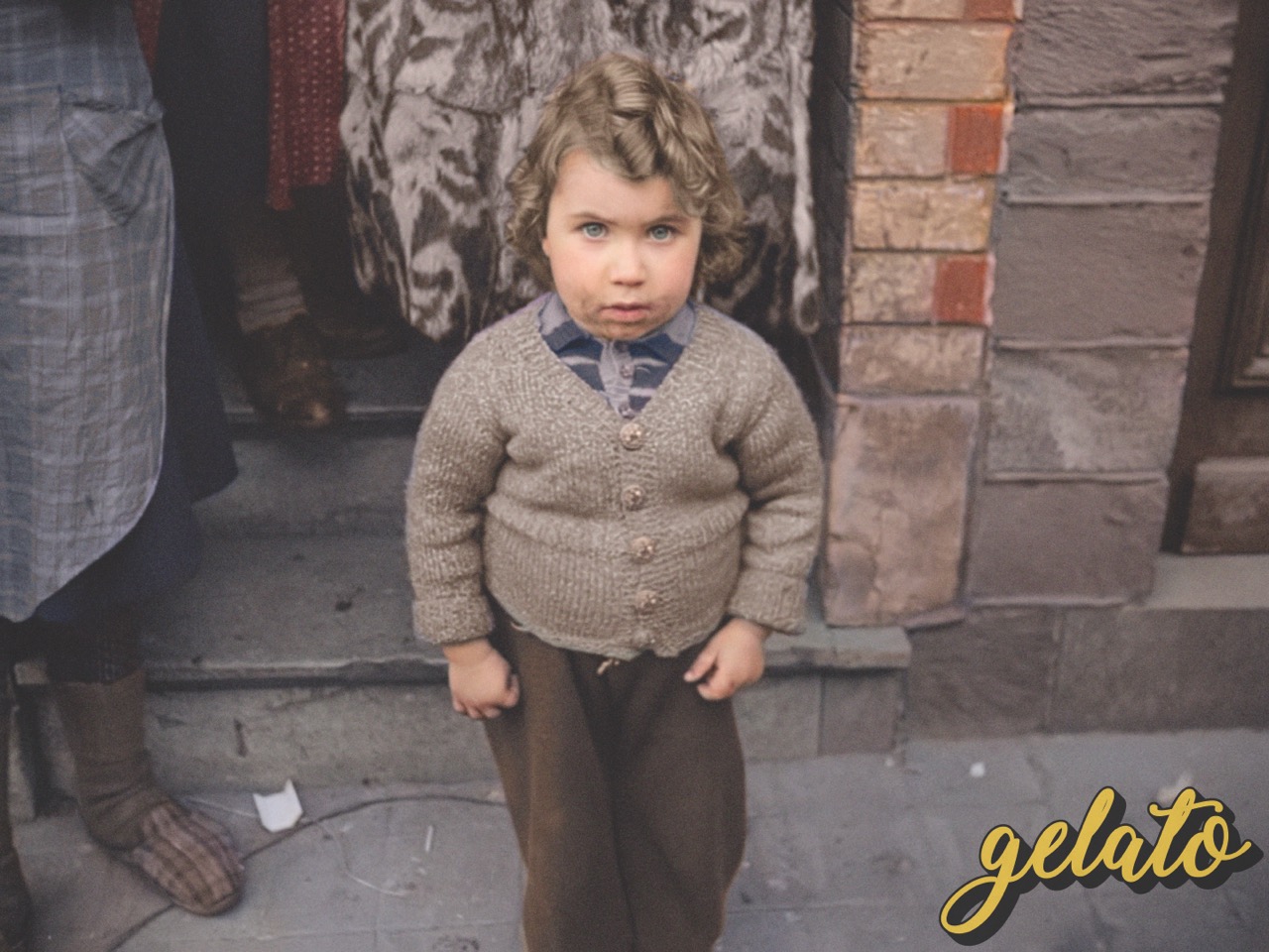 A black-and-white image of a small child standing next to a brick wall looking up innocently