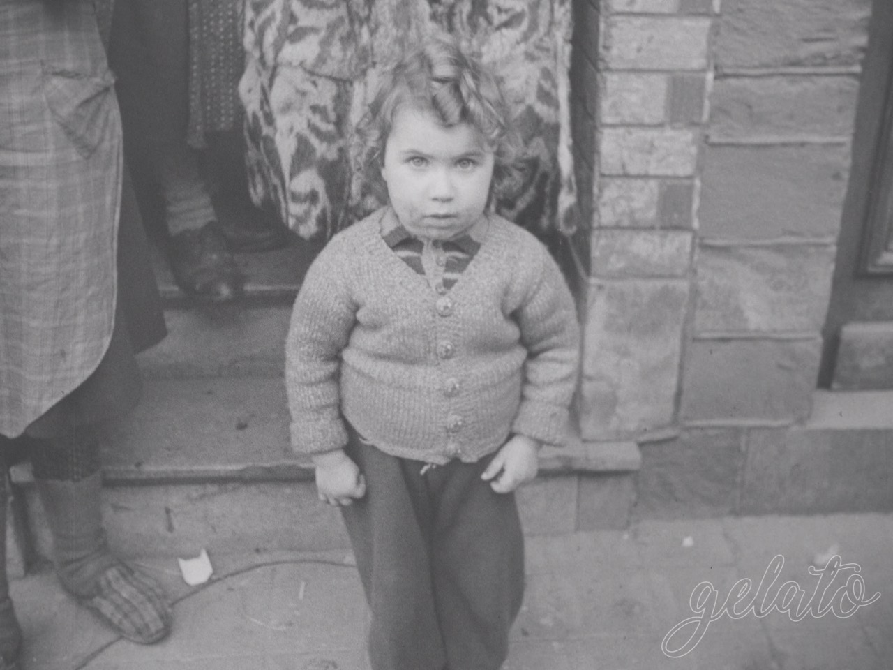 A black-and-white image of a small child standing next to a brick wall looking up innocently