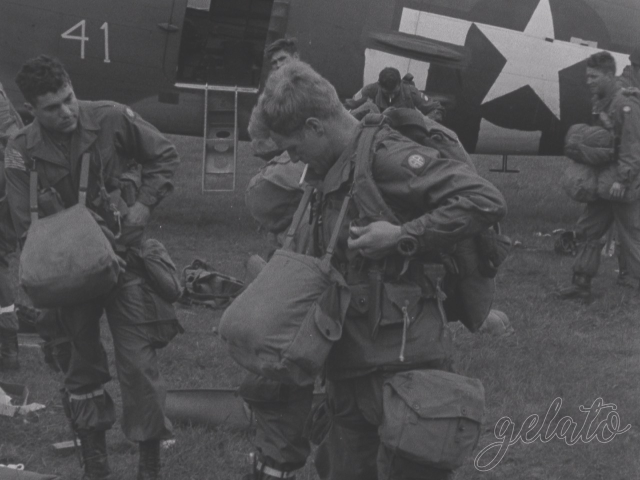 A faded photo of a soldier carrying multiple bags around his neck. He stands outside next to a plane