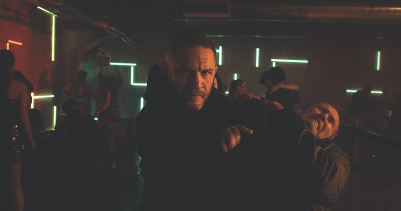 A dimly lit room with a man lit in red throttling another man in pain