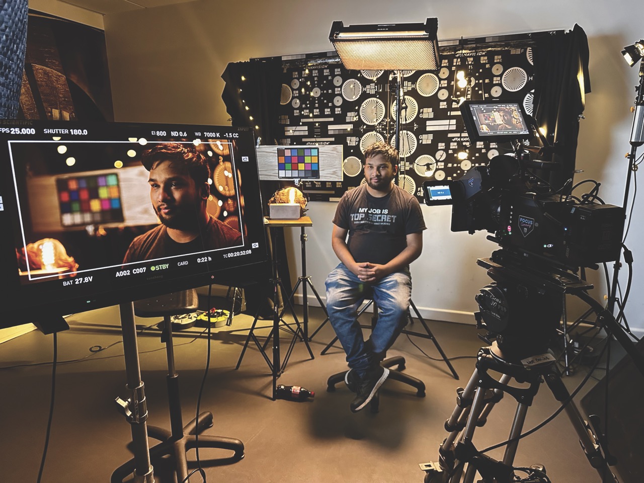 A man sitting in a studio while a camera films him and a monitor in the foreground shows his face