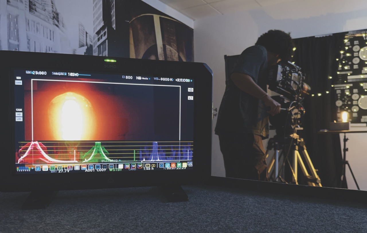 A dim room with a monitor showing light and a graph in the foreground, and a man adjusting a camera in the background