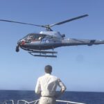 A man wearing white stands at the front of a boat in the sea. He looks at a low flying helicopter