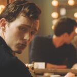 A young man with styled hair looks over his shoulder. He is reflected in the glowing mirror in front of him