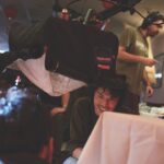 An actor with a devilish look sits by a table while two people behind and a person in front operate the technology