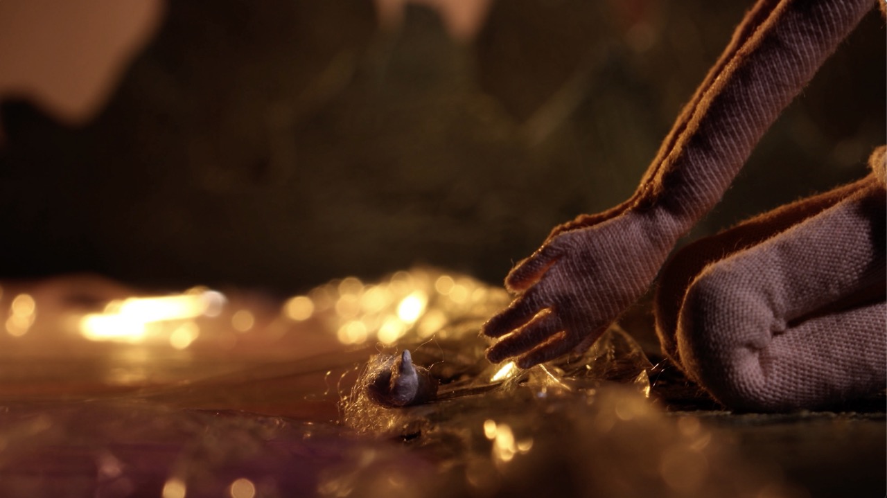 A handmade figurines hands picking something up from the wooden floor strewn with fairy lights