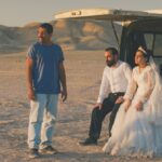 A bride and groom sit in the back of an open van while a man in a t-shirt looks into the distance beside them. The van is parked in a desert terrain