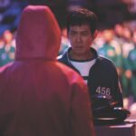 A man in a green tracksuit stands opposite a man wearing a pink hood. A crowd stands behind the man in the tracksuit