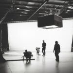 A black-and-white photo of a large warehouse. A group of people set up rigs and lighting technology with a large white sheet hanging behind them