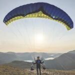 A person stands on the edge of a mountain with a mountainous terrain in the distance. They are attached to a yellow and blue parachute