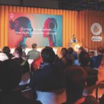 The IBC conference with a man speaking at a stand at the front of an orange room