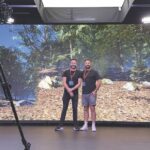 Two men stand close together in a virtual production studio. The LED screen behind shows a forest and there is a camera in front