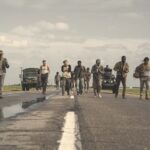 A group of young boys walk down a road followed by military trucks