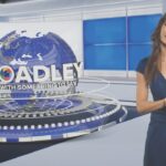 A woman stands in a news broadcasting room in front of Broadley's logo
