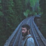 A man with a beard stands in the middle of train tracks bordered by trees