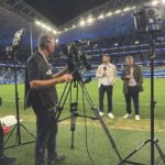 Two presenters stand on a football pitch with microphones while a man films them on a professional camera at nighttime