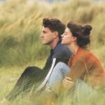 A man and woman sitting in a grassy field on the side of a hill