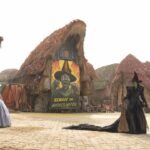 A still from Wicked showing two women facing each other dressed as a witch and fairy in the middle of a mythical town