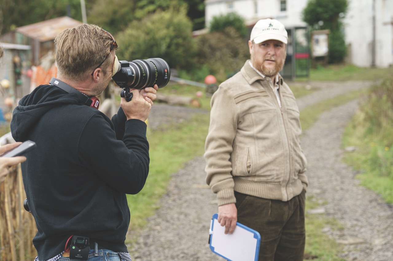 A still from The Ballad of Wallis Island showing a man wearing a cap being filmed by a man with a camera