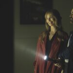 A still from A Big Bold Beautiful Journey where a man and woman stand smiling in a dim room with a torch