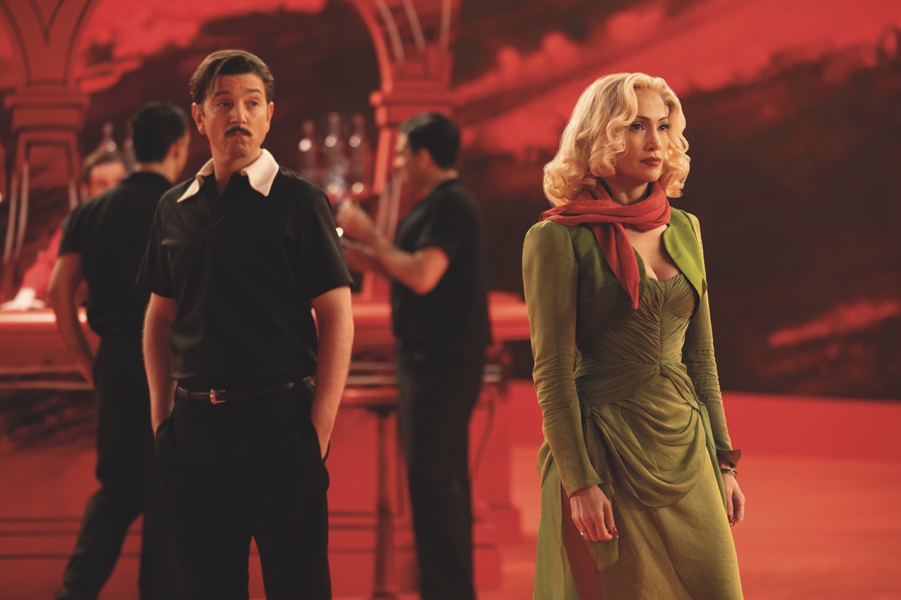 The saturated Technicolor of Molina’s imagined world is contrasted with the cold, claustrophobic prison setting A still from Kiss of the Spider Woman showing a woman in a green dress on a red film set with a man looking at her