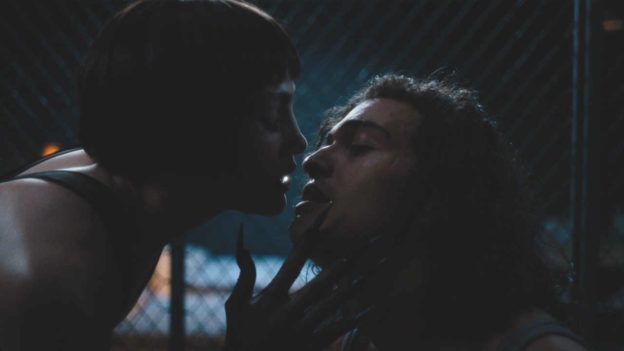 The saturated Technicolor of Molina’s imagined world is contrasted with the cold, claustrophobic prison setting A dimly lit still from Kiss of the Spider Woman showing a person leaning close into the face of another person