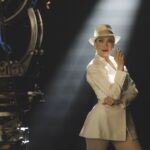 A still from Kiss of the Spider Woman showing a woman in a white suit and hat posing with a cigarette in a spotlight