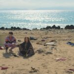 A man and a woman sit on a deserted beach with debris around them