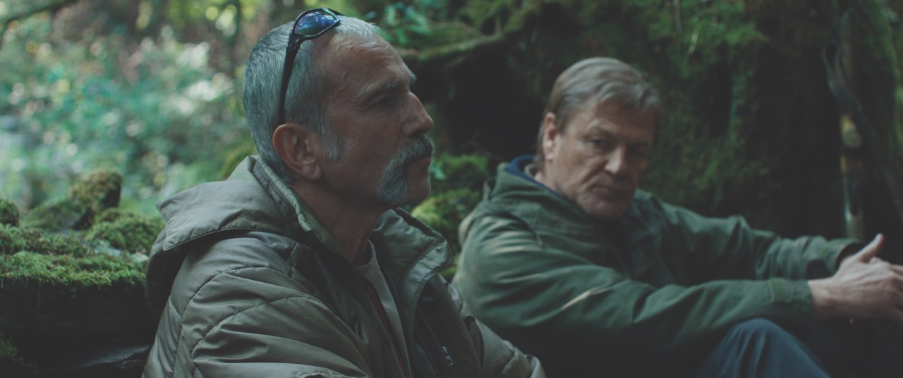 A still from Anemone showing two men in khaki coats, sitting in a wood