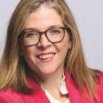 A professional photo of a smiling woman in a suit with glasses, against a grey background