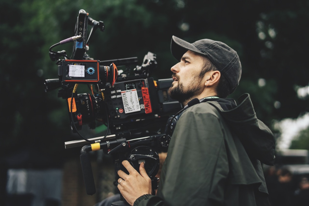 A person wearing a cap and operating a large camera from RED outside on a street