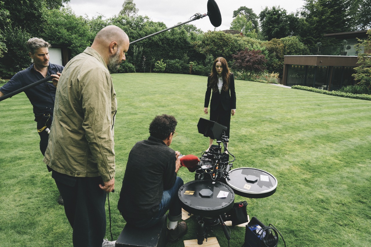 An actress in a black skirt and holding an umbrella in a field, surrounded by a film crew
