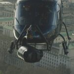 A helicopter with a large camera attached flying over a city