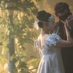 A man and woman in period dress at a masquerade ball. The man kisses the woman's wrist, illuminated in soft light and surrounded by foliage