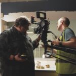 A person holding a professional camera while another person makes adjustments in a kitchen area