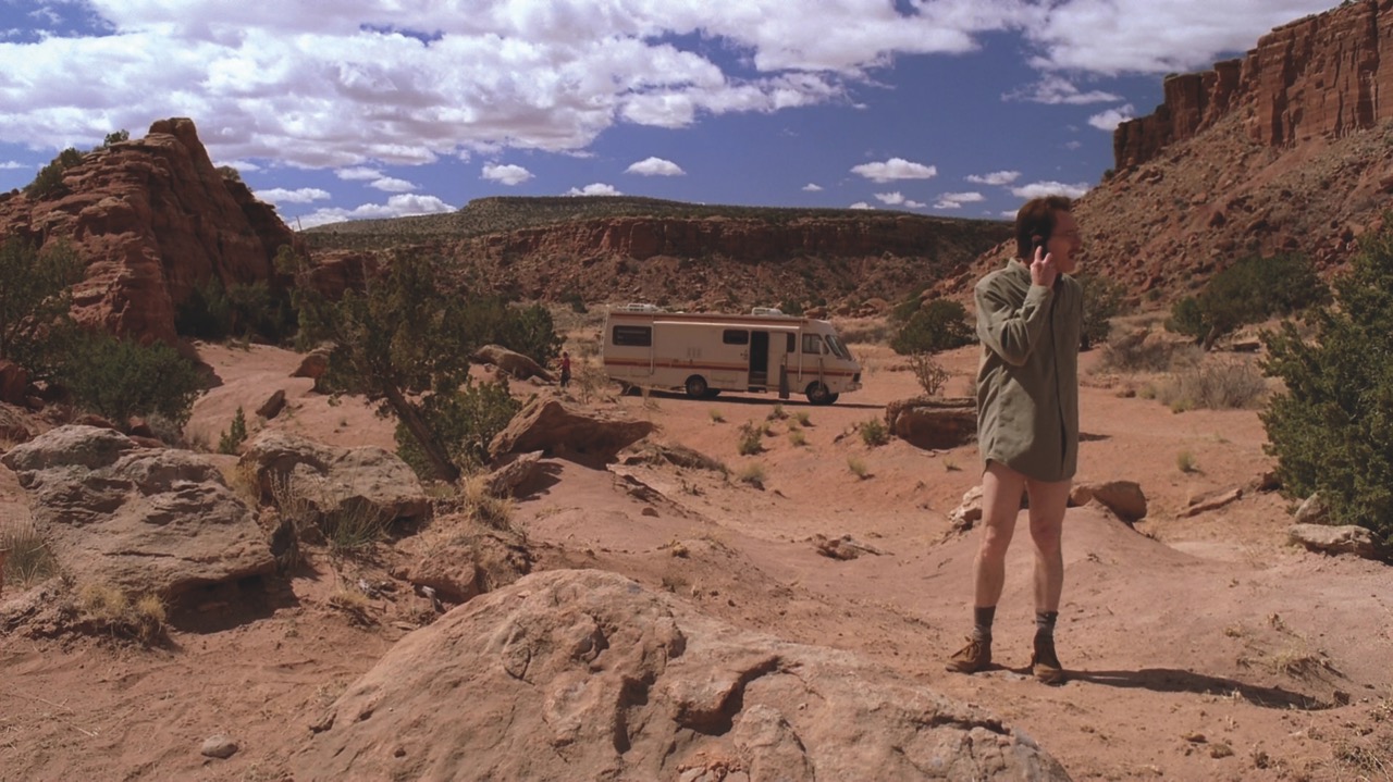 A man wearing shorts and standing in a desert environment with a caravan in the background